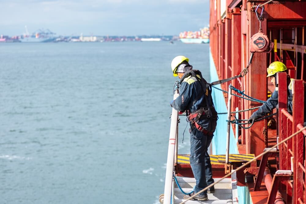 Maritime worker wearing safety gear and harness performing maintenance on a cargo ship.