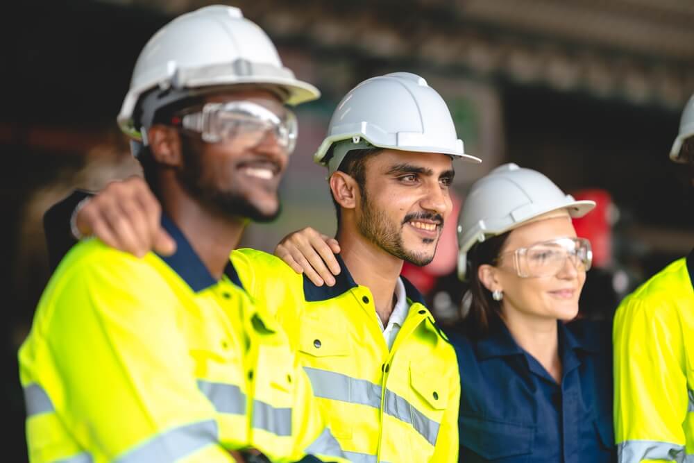 Group of industrial workers standing with arms around each other for a photo. They are wearing hard hats and safety jackets.