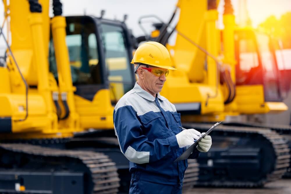 Industrial worker male senior in helmet holds tablet computer on background of production of excavator factory