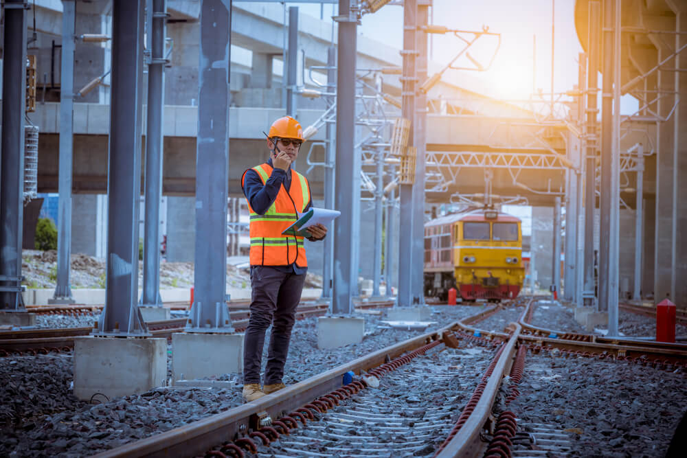 Rail worker standing on rail tracks talking into a radio with a train in the background.