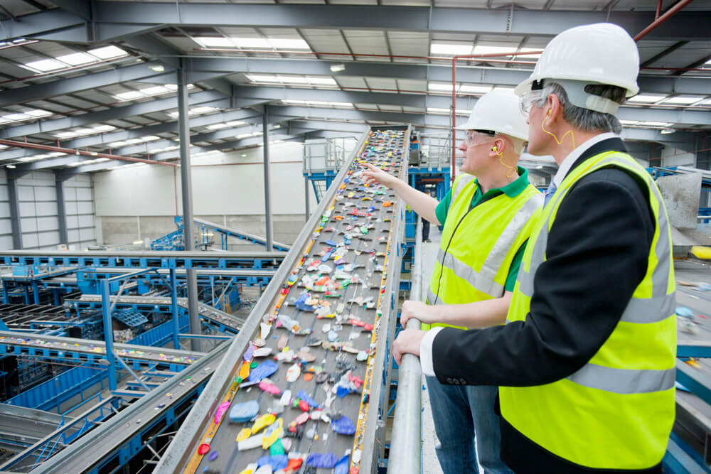 Two employees in protective jackets and hard hats standing over a conveyor line of waste being sorted.