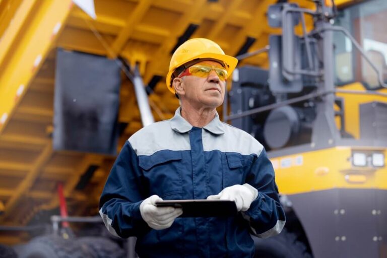 Man in safety glasses, gloves and hard hat holding a tablet device in front of mining or quarrying machinery.