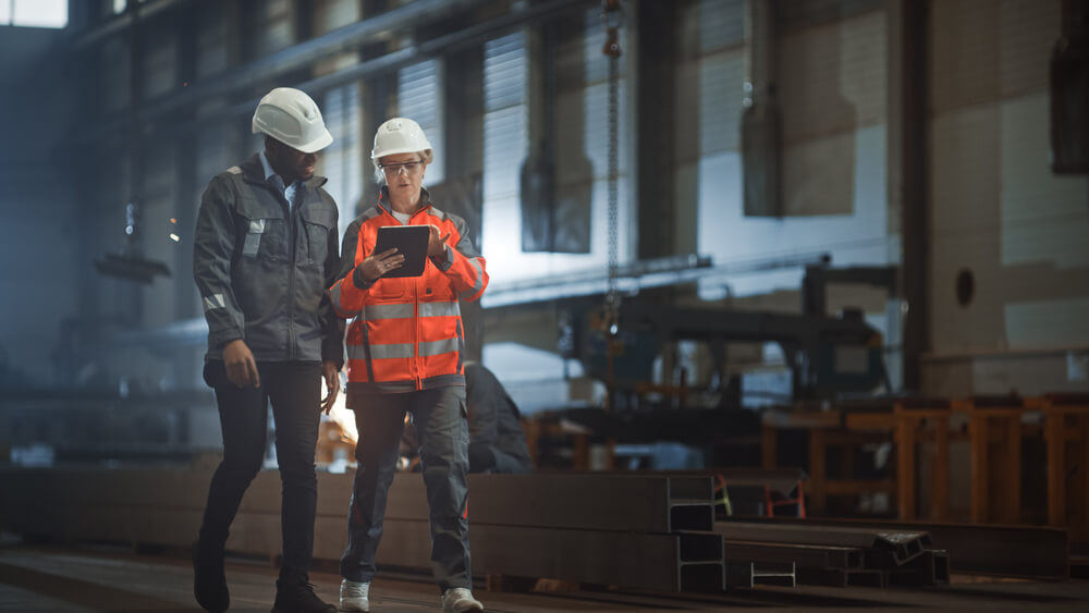 Two industrial engineers walking from industrial manufacturing operational plant or factory. Both are looking at a tablet computer.
