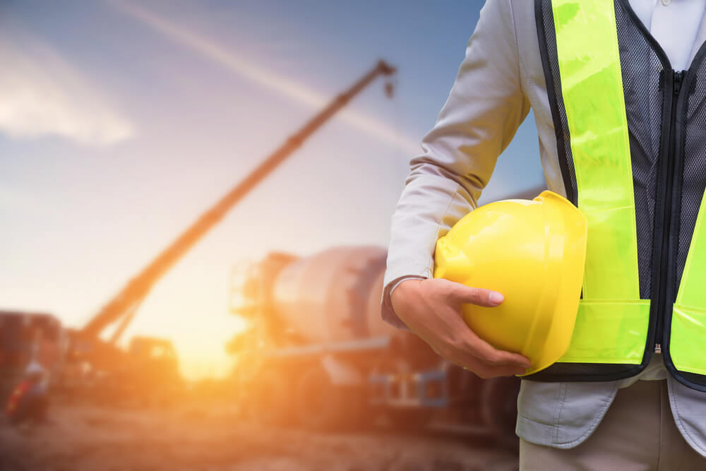 Man holding a hard hat and wearing a high visibility jacket with a crane and heavy machinery in the background.
